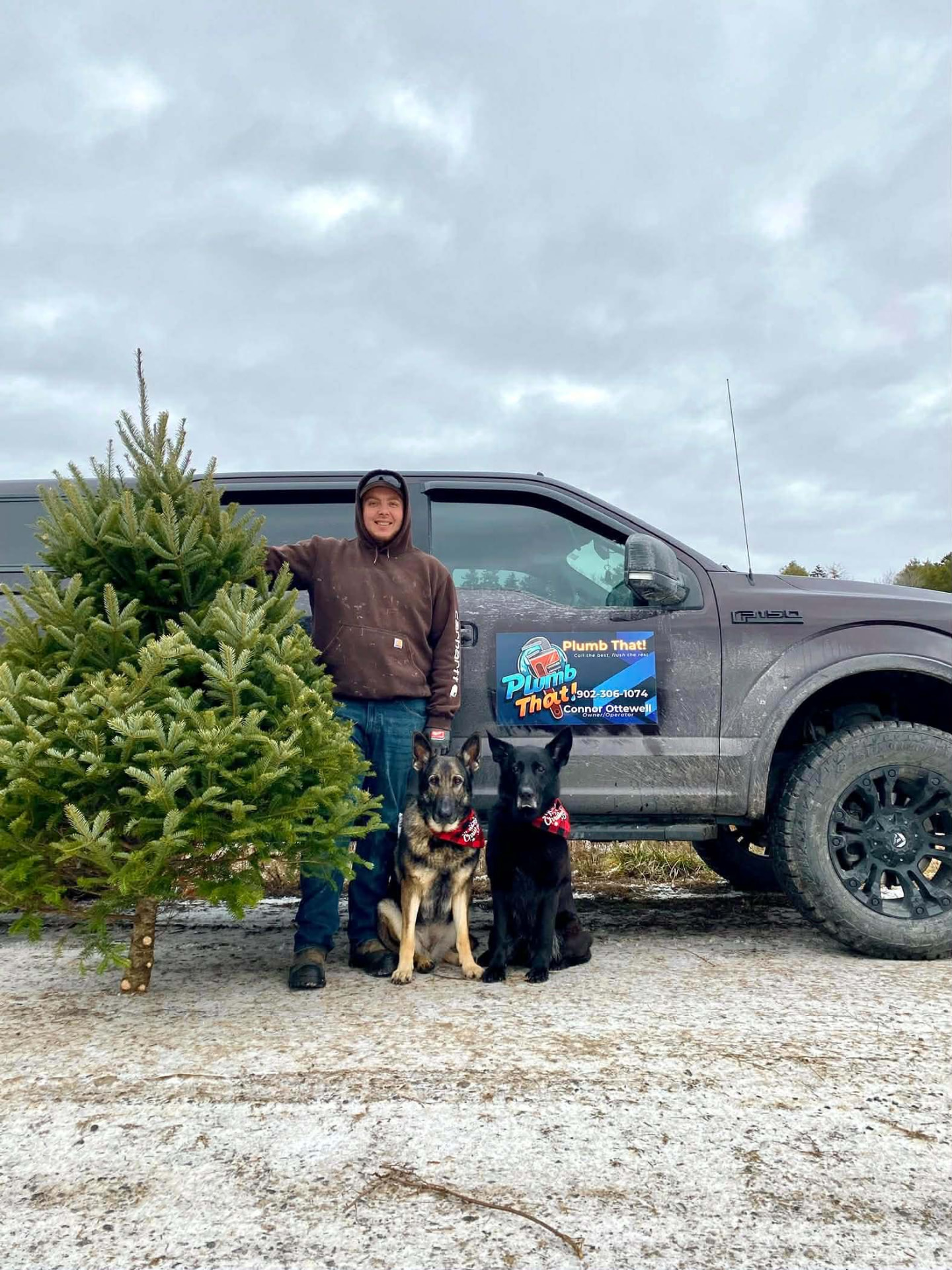 Connor Ottewell standing with his two dogs beside a Christmas tree, in front of his Plumb That! truck